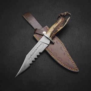 May include: A fixed-blade knife with a textured antler handle and a silver-toned steel blade featuring a serrated edge and engraved script. The knife rests on a brown leather sheath with visible stitching and a snap closure button.