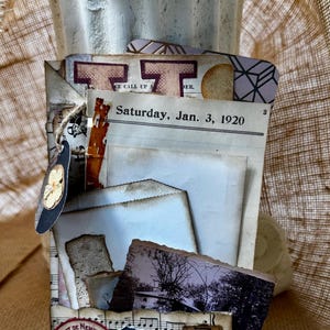 May include: A collage of vintage ephemera, including newspaper clippings, photographs, and sheet music. The text "Saturday, Jan. 3, 1920" is visible. A small blue teacup and a stamp are also included in the design. The collage is tied together with twine.