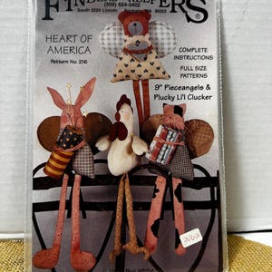 May include: A package for a craft pattern titled "Heart of America" featuring three fabric angel figures: a bunny, a chicken, and a cat. The pattern includes full-size patterns and complete instructions. The figures are approximately 9 inches tall.
