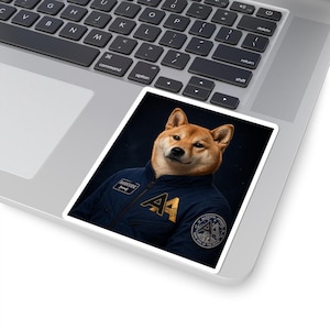 May include: A sticker featuring a Shiba Inu dog wearing a dark blue jacket with gold accents and the text "AA". The sticker has a dark background and is placed on a laptop.