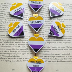 May include: Heart-shaped bookmarks with yellow, white, purple, and black designs. Each bookmark features a different phrase, including "PROUD," "NONBINARY BABY," "THEY/THEM," "LOVE IS LOVE," "SHOW YOUR COLOURS!," "IN MY READING ERA," and "SORRY, I'M ALL BOOKED."