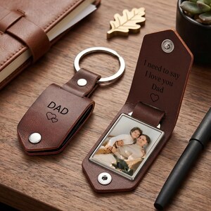 Op de afbeelding: Twee bruine leren sleutelhangers met zilverkleurige metalen ringen. De ene is gesloten en gegraveerd met DAD en twee hartjes. De andere is open en toont een vierkante foto van twee personen en de tekst I need to say I love you Dad.
