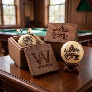 May include: Two cream-coloured billiard balls and two square wooden boxes. One ball features the name Wilson and a number 8 graphic. The other ball displays the name Anderson with a large letter A. The wooden boxes are engraved with matching designs.