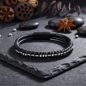 May include: A black leather bracelet featuring a double-strand design with a matte black magnetic clasp. The leather cord is wrapped with silver-toned metal beads and thin silver-toned metal bars arranged in a repeating pattern along the front.