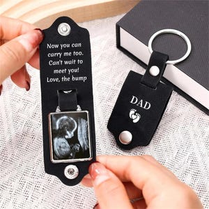 Peut inclure: Deux porte-clés en cuir noir avec des anneaux en métal argenté et des boutons-pression. L'un est ouvert, montrant une échographie en noir et blanc et le texte : Now you can carry me too. Can't wait to meet you! Love, the bump. L'autre est fermé, avec le mot DAD et deux empreintes de pieds de bébé.