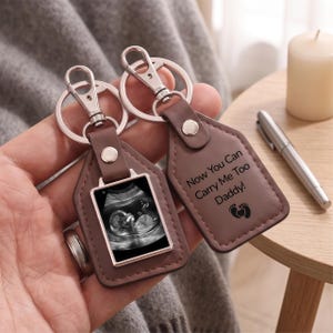 Op de afbeelding: Twee bruine lederen sleutelhangers met zilverkleurige metalen hardware. Eén sleutelhanger bevat een zwart-wit echografie van een foetus. De andere sleutelhanger is gegraveerd met de tekst "Now You Can Carry Me Too Daddy!" en een icoon van voetafdrukjes.