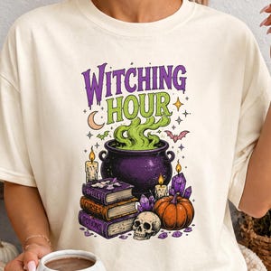 May include: An ivory t-shirt featuring a graphic print with the text "WITCHING HOUR" in purple and green. The design includes a purple cauldron with green smoke, a stack of books labeled "SPELLS," "CURSES," and "POTIONS," a skull, and a pumpkin.