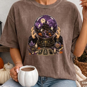 May include: An espresso-colored t-shirt featuring a graphic print of a crystal ball held by hands, displaying a haunted castle, ghosts, and pumpkins. The design includes tarot cards, a book, and candles. The shirt has a crew neck and short sleeves.