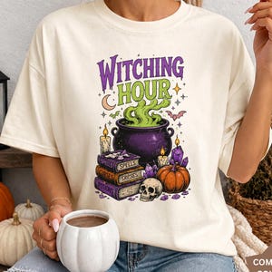 May include: An ivory t-shirt featuring a graphic print with the text "WITCHING HOUR" in purple and green. The design includes a purple cauldron with green smoke, a stack of books labeled "SPELLS," "CURSES," and "POTIONS," a skull, and a pumpkin.