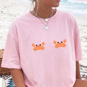 May include: A light pink short-sleeved t-shirt featuring two small, orange cartoon crab graphics positioned on the chest area. The shirt has a crew neckline and a relaxed fit. The word Blossom is printed in a small pink box in the top corner.