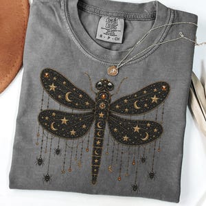 May include: A pepper-coloured crew neck t-shirt featuring a graphic of a large dragonfly with black wings adorned with gold stars, crescent moons, and dangling chains with small spiders. The shirt has a Comfort Colors label at the inner neckline.