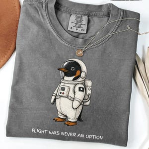 May include: A charcoal grey crew neck t-shirt featuring a graphic of a penguin wearing a white astronaut space suit. Below the graphic, the text reads FLIGHT WAS NEVER AN OPTION. The shirt has a Comfort Colors label inside the collar.
