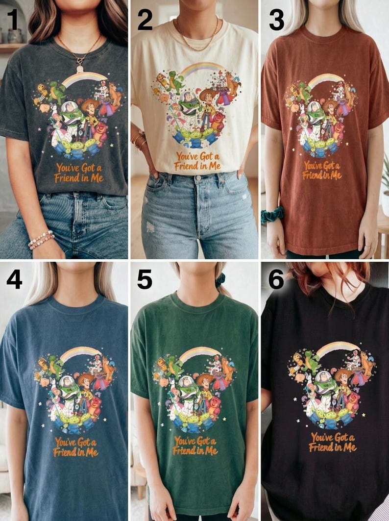 K&ouml;nnte beinhalten: Sechs verschiedenfarbige T-Shirts mit einer Grafik von Toy Story-Charakteren in Form von Mickey Mouse-Ohren, einem Regenbogen und dem Text "You've Got a Friend in Me". Die Farben sind Anthrazit, Creme, Rost, Blau, Gr&uuml;n und Schwarz.
