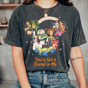 K&ouml;nnte beinhalten: Dunkelgraues T-Shirt mit einer farbenfrohen Grafik von Toy Story-Charakteren, darunter Woody, Buzz Lightyear und die kleinen gr&uuml;nen M&auml;nnchen. Der Text "You've Got a Friend in Me" ist in Orange gedruckt. Das T-Shirt wird von einer Person getragen.