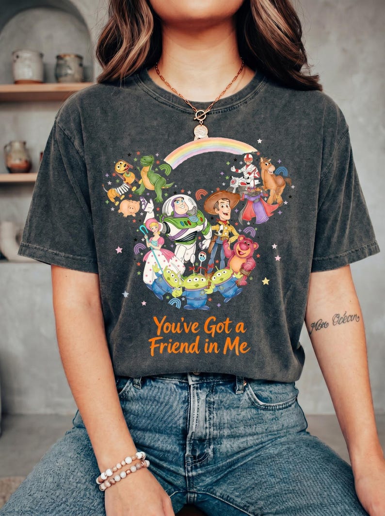K&ouml;nnte beinhalten: Dunkelgraues T-Shirt mit einer farbenfrohen Grafik von Toy Story-Charakteren, darunter Woody, Buzz Lightyear und die kleinen gr&uuml;nen M&auml;nnchen. Der Text "You've Got a Friend in Me" ist in Orange gedruckt. Das T-Shirt wird von einer Person getragen.