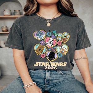 Puede incluir: Camiseta gris oscuro con un dise&ntilde;o gr&aacute;fico colorido de personajes de Star Wars dentro de marcos en forma de coraz&oacute;n, rodeados de flores. El texto "STAR WARS 2026" est&aacute; impreso debajo. La camiseta es de material suave.