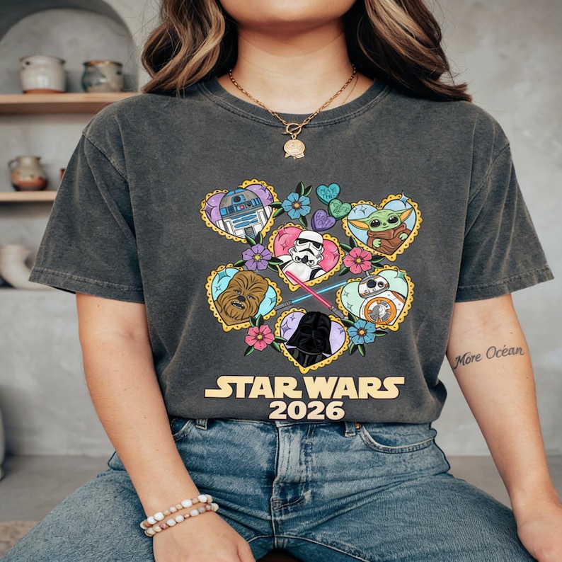 Puede incluir: Camiseta gris oscuro con un dise&ntilde;o gr&aacute;fico colorido de personajes de Star Wars dentro de marcos en forma de coraz&oacute;n, rodeados de flores. El texto "STAR WARS 2026" est&aacute; impreso debajo. La camiseta es de material suave.