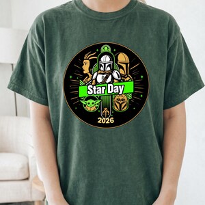 May include: Dark green t-shirt featuring a circular graphic with Star Wars characters and the text "Star Day 2026". The design includes images of a Mandalorian helmet, Yoda, and other characters. The shirt is a comfortable, casual style.