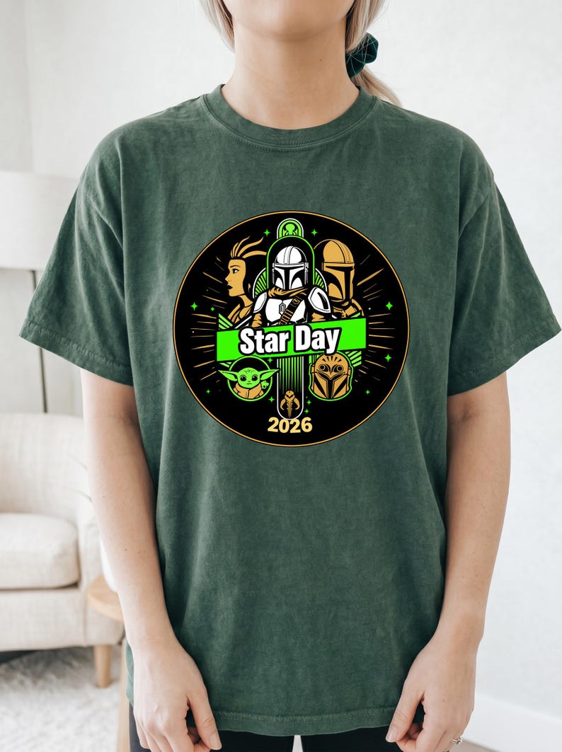 May include: Dark green t-shirt featuring a circular graphic with Star Wars characters and the text "Star Day 2026". The design includes images of a Mandalorian helmet, Yoda, and other characters. The shirt is a comfortable, casual style.