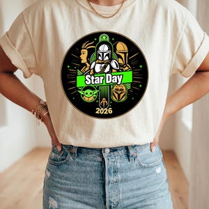 May include: Cream-colored t-shirt featuring a circular graphic with Star Wars characters and the text "Star Day 2026". The design includes images of a Mandalorian, a Jedi, and other characters from the Star Wars franchise.