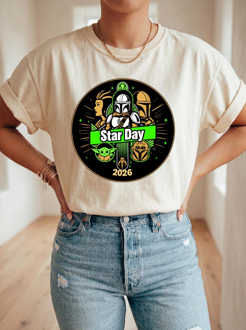 May include: Cream-colored t-shirt featuring a circular graphic with Star Wars characters and the text "Star Day 2026". The design includes images of a Mandalorian, a Jedi, and other characters from the Star Wars franchise.