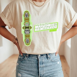 Pu&ograve; includere: T-shirt color crema con grafica verde neon con personaggi di Star Wars e la scritta "STAR WARS DAY MAY THE 4TH BE WITH YOU 2026". La maglietta &egrave; abbinata a jeans azzurri.