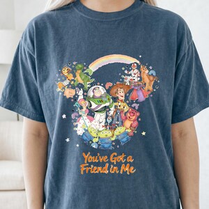 K&ouml;nnte beinhalten: Marineblaues T-Shirt mit einer farbenfrohen Grafik von Toy Story-Charakteren, angeordnet in Form eines Mickey Mouse-Kopfes. Der Text "You've Got a Friend in Me" ist orangefarben gedruckt.