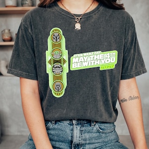 Pu&ograve; includere: T-shirt grigio scuro con grafica verde neon con personaggi di Star Wars e la scritta "STAR WARS DAY MAY THE 4TH BE WITH YOU 2026". La maglietta ha girocollo e maniche corte.
