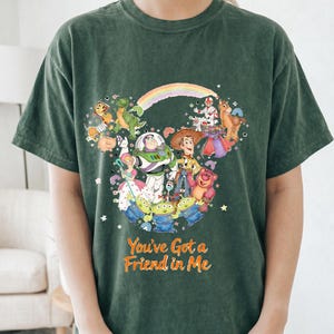 K&ouml;nnte beinhalten: Dunkelgr&uuml;nes T-Shirt mit einer farbenfrohen Illustration von Toy Story-Charakteren, angeordnet in Form eines Mickey Mouse-Kopfes. Der Text "You've Got a Friend in Me" ist darunter aufgedruckt. Das Shirt besteht aus einem weichen Material.