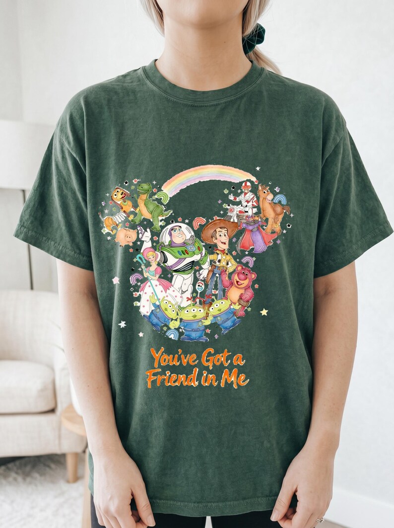 K&ouml;nnte beinhalten: Dunkelgr&uuml;nes T-Shirt mit einer farbenfrohen Illustration von Toy Story-Charakteren, angeordnet in Form eines Mickey Mouse-Kopfes. Der Text "You've Got a Friend in Me" ist darunter aufgedruckt. Das Shirt besteht aus einem weichen Material.