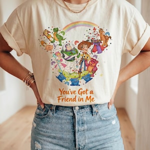 K&ouml;nnte beinhalten: Cremefarbenes T-Shirt mit einem farbenfrohen Grafikdesign von Toy Story-Charakteren, darunter Woody und Buzz Lightyear. Das Design zeigt einen Regenbogen und den Text "You've Got a Friend in Me". Die &Auml;rmel sind hochgekrempelt.