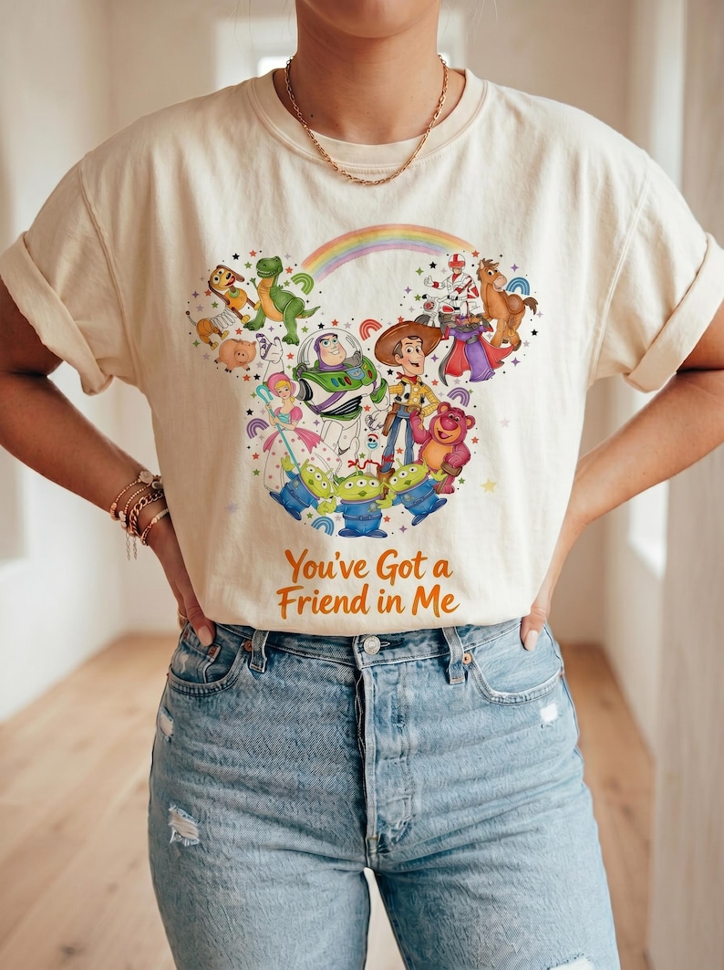 K&ouml;nnte beinhalten: Cremefarbenes T-Shirt mit einem farbenfrohen Grafikdesign von Toy Story-Charakteren, darunter Woody und Buzz Lightyear. Das Design zeigt einen Regenbogen und den Text "You've Got a Friend in Me". Die &Auml;rmel sind hochgekrempelt.