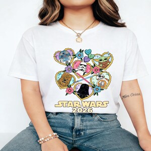 Puede incluir: Camiseta blanca con un dise&ntilde;o gr&aacute;fico colorido de personajes de Star Wars en marcos con forma de coraz&oacute;n y detalles florales. El dise&ntilde;o incluye a R2-D2, Chewbacca, Darth Vader y Baby Yoda. El texto "STAR WARS 2026" est&aacute; impreso debajo.