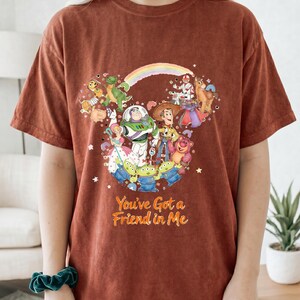 K&ouml;nnte beinhalten: Rostfarbenes T-Shirt mit einem farbenfrohen Grafikdesign von Toy Story-Charakteren, darunter Buzz Lightyear und Woody. Der Text "You've Got a Friend in Me" ist darunter gedruckt. Das T-Shirt hat kurze &Auml;rmel.