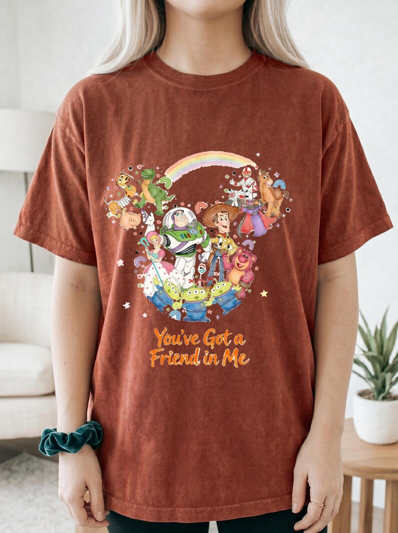 K&ouml;nnte beinhalten: Rostfarbenes T-Shirt mit einem farbenfrohen Grafikdesign von Toy Story-Charakteren, darunter Buzz Lightyear und Woody. Der Text "You've Got a Friend in Me" ist darunter gedruckt. Das T-Shirt hat kurze &Auml;rmel.
