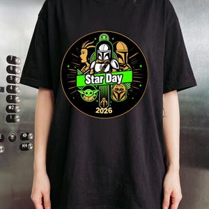 May include: Black t-shirt featuring a circular graphic with characters and the text "Star Day 2026". The design includes images of a Mandalorian, Grogu, and other characters from the Star Wars franchise. The graphic is in green, white, and gold.