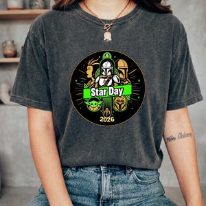 May include: Dark gray t-shirt featuring a circular graphic with characters and the text "Star Day 2026". The design includes green and yellow accents, and images of iconic characters from a popular science fiction franchise.