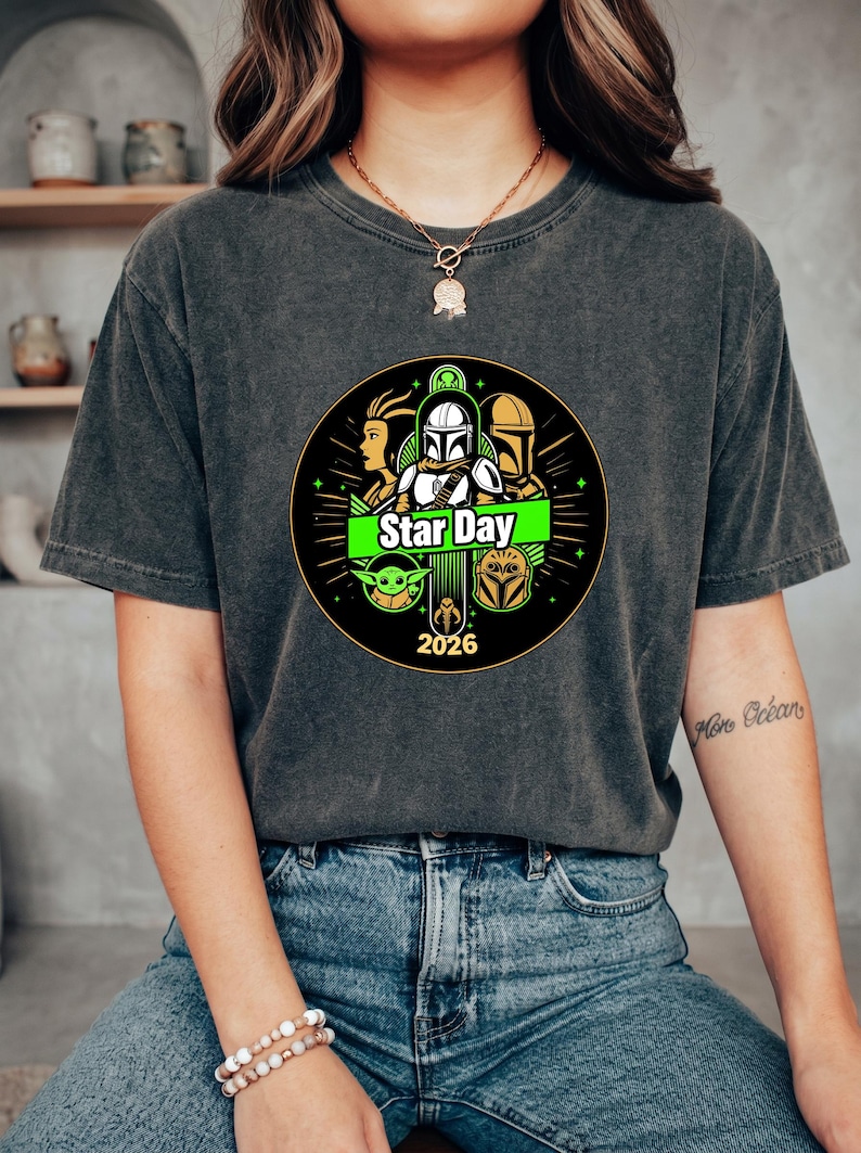 May include: Dark gray t-shirt featuring a circular graphic with characters and the text "Star Day 2026". The design includes green and yellow accents, and images of iconic characters from a popular science fiction franchise.