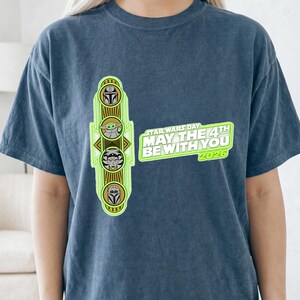 Pu&ograve; includere: T-shirt blu con grafica verde neon con personaggi di Star Wars e la scritta "STAR WARS DAY MAY THE 4TH BE WITH YOU 2026". La grafica include immagini dei personaggi del franchise di Star Wars.