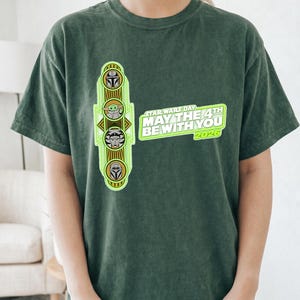 Pu&ograve; includere: T-shirt verde scuro con un design del Star Wars Day. Il design include illustrazioni di personaggi e il testo "MAY THE 4TH BE WITH YOU 2026". La maglietta &egrave; realizzata in un materiale morbido.