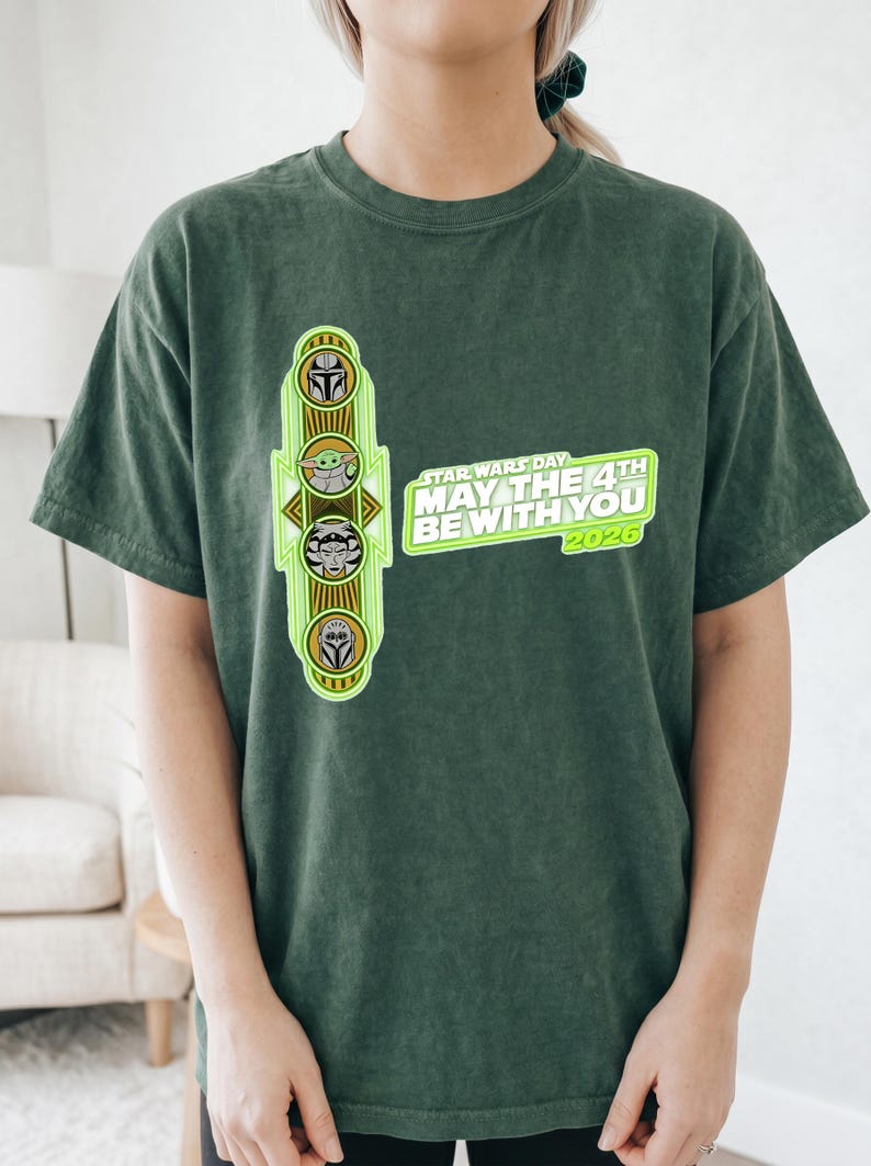 Pu&ograve; includere: T-shirt verde scuro con un design del Star Wars Day. Il design include illustrazioni di personaggi e il testo "MAY THE 4TH BE WITH YOU 2026". La maglietta &egrave; realizzata in un materiale morbido.