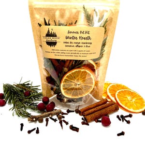May include: A clear bag labeled "Simmer Pot Kit Winter Hearth" filled with dried orange slices, cinnamon sticks, cloves, and other botanicals. The kit includes instructions for simmering with water. Sprigs of rosemary and cranberries are scattered around the bag.