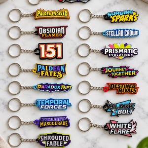 May include: A collection of seventeen colorful rubber keychains featuring various Pokémon trading card set logos including Scarlet & Violet, Paldea Evolved, Obsidian Flames, 151, Paldean Fates, Paradox Rift, Temporal Forces, Twilight Masquerade, and Shrouded Fable.
