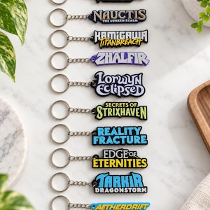 Puede incluir: Una colección de diez llaveros de goma rectangulares dispuestos verticalmente, cada uno con un anillo y cadena de metal plateado. Los diseños muestran varios logotipos y textos temáticos de Magic the Gathering, incluyendo Nauctis, Kamigawa, Zhalfir, Lorwyn, Strixhaven, Reality Fracture, Edge of Eternities, Tarkir y Aetherdrift.