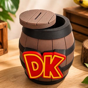 Könnte beinhalten: Eine braune, fassförmige Spardose mit schwarzen horizontalen Bändern und einem abnehmbaren runden Deckel mit Münzschlitz. Auf der Vorderseite des Fasses befinden sich die Buchstaben DK in kräftigem Rot mit gelbem Rand, ähnlich einem Holzfass.