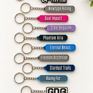 May include: A collection of ten keychains featuring various colors and text including Gundam Card Game, Newtype Rising, Dual Impact, Steel Requiem, Phantom Aria, Eternal Nexus, Freedom Ascension, Stardust Trails, Blazing Fist, and GDB.