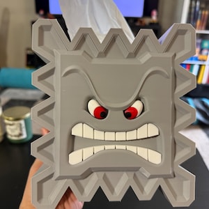 Puede incluir: Una caja de pañuelos gris con forma de Thwomp de la serie de videojuegos Super Mario. La caja presenta un diseño cuadrado con un borde dentado, una cara fruncida con ojos rojos y dientes blancos. Se ven pañuelos blancos.