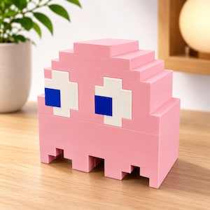 May include: A pink pixelated ghost figure constructed from interlocking plastic building blocks. The figure features a rounded top, two large white eyes with centered blue square pupils, and a jagged bottom edge with three distinct rectangular feet.
