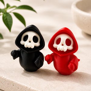 May include: Two small figurines featuring white skulls with hollow eye sockets, each wearing a hooded robe. One figurine has a black robe, and the other has a red robe. Both figures have rounded bodies and small arms extended forward in a pose.