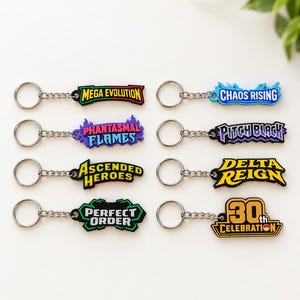 May include: Eight metal keychains with colorful rubberized tags featuring text: Mega Evolution, Chaos Rising, Phantasmal Flames, Pitch Black, Ascended Heroes, Delta Reign, Perfect Order, and 30th Celebration with a small circular icon.